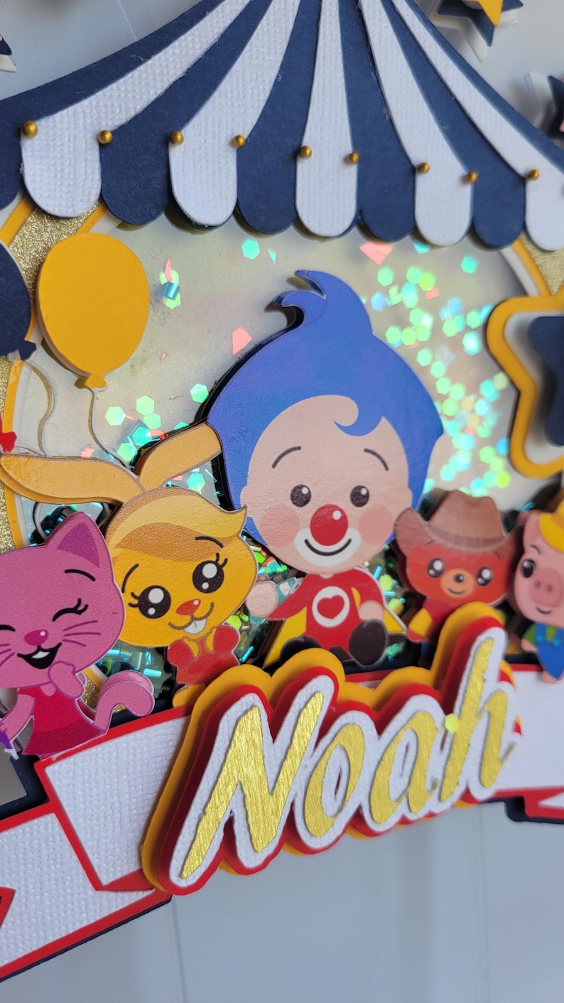 May include: A colorful, layered cake topper featuring cartoon characters and the name "Noah" in gold lettering. The design includes a circus tent, balloons, and various animal figures, creating a festive and celebratory theme.