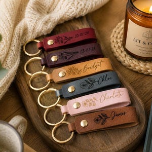 May include: A set of six leather keychains in various colors including red, dark purple, tan, dark gray, light pink, and brown. Each keychain features a gold-toned metal ring, a gold-toned snap button, and custom laser-engraved floral designs and names.