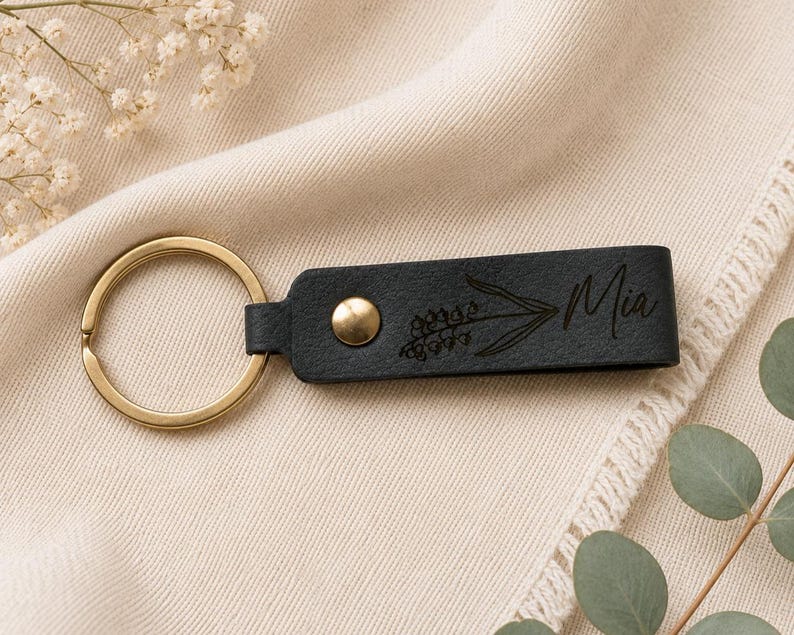 Close-up of a personalized leather keychain featuring a custom engraved name and birth flower. The detailed botanical engraving and smooth leather texture are highlighted with brass hardware.