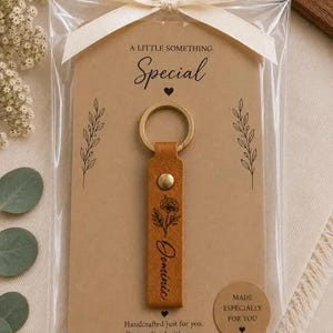 May include: A tan leather keychain with a gold metal ring and rivet, featuring a black floral illustration and the name Dominic. It is attached to a tan card with the text A LITTLE SOMETHING Special, Made especially for you, and a white ribbon.