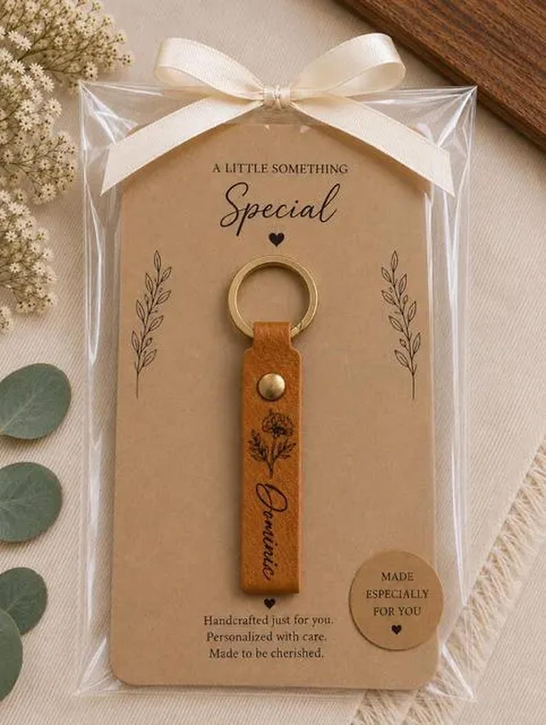 May include: A tan leather keychain with a gold metal ring and rivet, featuring a black floral illustration and the name Dominic. It is attached to a tan card with the text A LITTLE SOMETHING Special, Made especially for you, and a white ribbon.