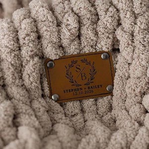 May include: Close-up of a chunky knit blanket in a neutral beige color. A brown leather tag is attached, featuring the initials "S" and "B" within a laurel wreath design, along with the names "Stephen + Bailey" and the date "12.10.2026".