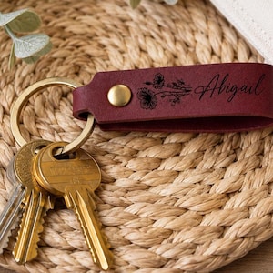 May include: A deep red leather keychain featuring a gold-toned metal rivet and a gold-toned split ring holding three metal keys. The leather strap is engraved with a black floral illustration and the name Abigail in a cursive script font.