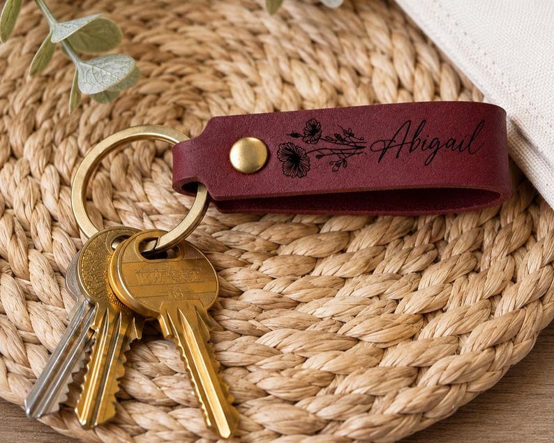 May include: A deep red leather keychain featuring a gold-toned metal rivet and a gold-toned split ring holding three metal keys. The leather strap is engraved with a black floral illustration and the name Abigail in a cursive script font.