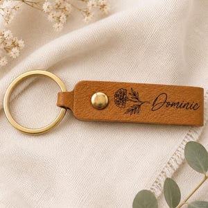 Custom engraved birth flower leather keychain styled on textured cream fabric with greenery and dried flowers. The botanical engraving and brass hardware create a timeless handcrafted keepsake.
