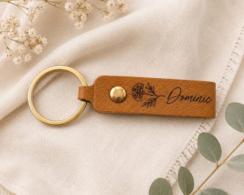 Custom engraved birth flower leather keychain styled on textured cream fabric with greenery and dried flowers. The botanical engraving and brass hardware create a timeless handcrafted keepsake.