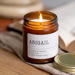 Puede incluir: Una vela encendida en un frasco de vidrio ámbar con una etiqueta blanca que muestra el texto ABIGAIL (noun) 1. rather be reading 2. dog lover 3. spicy romance girly. La vela tiene una tapa de metal dorado junto a ella sobre una tela texturizada.