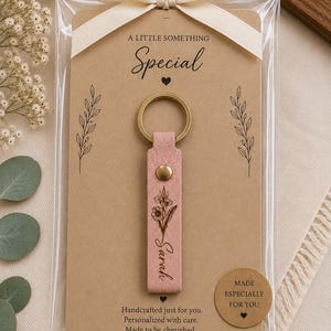 Engraved leather keychain featuring a personalized name surrounded by a delicate botanical birth flower design. Premium leather and brass hardware create an elegant everyday accessory.