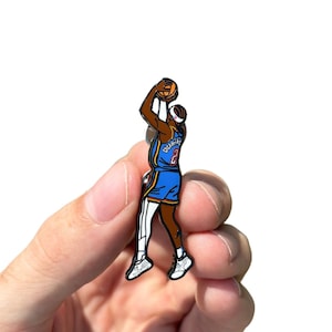 May include: A blue and white enamel pin of a basketball player with a basketball in hand. The pin is being held in a hand, ready to be worn.