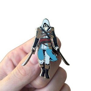 May include: Enamel pin featuring a stylized character in a hooded outfit, holding two swords. The character is depicted in shades of brown, blue, and white, with black boots and a red sash. The pin is held against a white background.