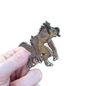 May include: An enamel pin featuring a brown and tan creature with horns, claws, and a spiky back. The pin is held against a white background. The creature has a detailed design with a menacing expression.