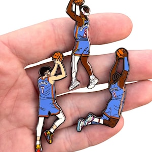 May include: Three enamel pins featuring basketball players in action. Each pin depicts a player in a blue and orange uniform, holding a basketball. The pins are detailed with white accents and black outlines, showcasing dynamic poses.