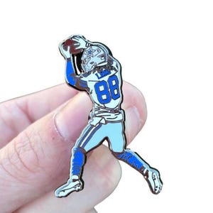 May include: A pin featuring a football player in a blue and white uniform catching a football. The player's jersey has the number 88 in blue. The pin is held by a person's fingers.