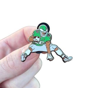 May include: A green and white enamel pin depicting a football player in action. The player is wearing a green helmet and jersey with the number 25. The pin is held in a hand against a white background.