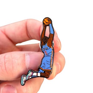 May include: Enamel pin featuring a basketball player in a blue jersey with the name "Williams" and the number "8". The player is mid-dunk, holding an orange basketball. The pin has a white background.