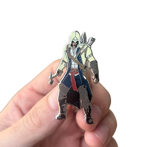 May include: A silver-colored enamel pin featuring a cartoon depiction of a video game character. The character is wearing a hooded cloak, a blue and red outfit, and holding a hidden blade. The pin has a detailed design with a black and white color scheme.