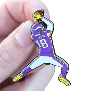 May include: Enamel pin featuring a football player in a purple and white uniform catching a football. The player's jersey has the number 18 and the name "Jefferson" on the back. The pin has yellow and black accents.