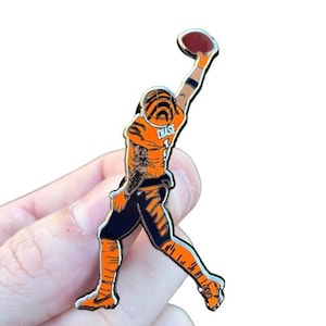 May include: A metal pin depicting a football player in an orange and black uniform, catching a brown football. The player's jersey has the name "CHASE" and the number "1". The pin is held by a hand.