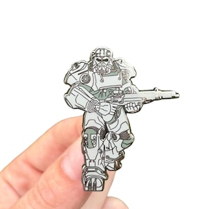 May include: A silver and gray enamel pin depicting a futuristic soldier in power armor, holding a rifle. The pin features detailed line art and a glossy finish, perfect for fans of science fiction and video games.