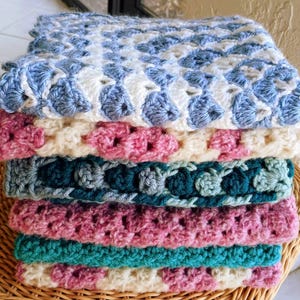 May include: A stack of colorful, handmade crochet blankets. The blankets feature a variety of colors, including blue, white, pink, teal, and cream. The crochet stitch creates a textured, patterned design. The blankets are neatly folded and stacked.