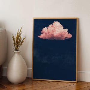 May include: A framed art print featuring a pink cloud against a dark blue background. The artwork is displayed on a wooden floor, leaning against a white wall. A beige sofa and a white vase with dried plants are also visible.