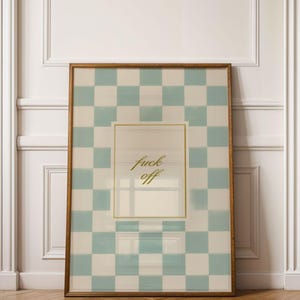 May include: A framed art print with a light brown wooden frame. The artwork features a checkered pattern in light blue and white, with the words "fuck off" in gold script. The print is leaning against a white wall with two white doors.