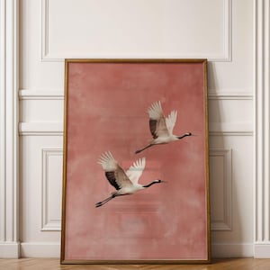 Op de afbeelding: Een ingelijste kunstdruk met twee kraanvogels in vlucht tegen een zachte, roze-rode achtergrond. De vogels hebben witte vleugels met zwarte accenten en de lijst is warm goudbruin. Het kunstwerk wordt getoond in een kamer met witte pilaren en een houten vloer.