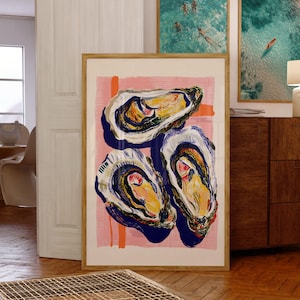 Op de afbeelding: Een ingelijste kunstdruk met drie gestileerde oesterillustraties. De oesters zijn afgebeeld met blauwe, gele en witte details tegen een roze achtergrond met oranje penseelstreken. Het kunstwerk wordt tentoongesteld in een kamer met andere ingelijste kunstwerken.