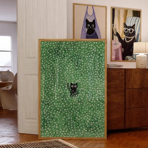 Op de afbeelding: Een ingelijste kunstdruk met een zwarte kat in een veld met witte bloemen tegen een groene achtergrond. Twee extra ingelijste kattenkunstdrukken zijn zichtbaar, een met een kat in een netzak en een andere met een kat met een zonnebril en een parelketting.