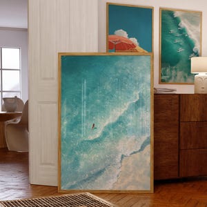 May include: A framed art print showing a swimmer in turquoise ocean water. The artwork is in a wooden frame and is displayed with two other framed prints, one with a beach umbrella and another with surfers.