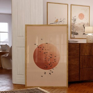 May include: A framed art print depicting a large orange sun with a flock of black birds in flight. The artwork is in a wooden frame and is displayed with other framed art pieces. The overall theme is nature and minimalist design.