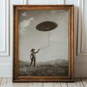 May include: Framed sepia-toned print featuring a cowboy roping a UFO in a desert landscape. The artwork has a vintage aesthetic, with a dark brown wooden frame. The sky is filled with clouds, and the ground is covered with sparse vegetation.