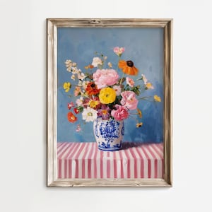 Op de afbeelding: Een ingelijst bloemstilleven schilderij. Een blauw-witte vaas bevat een boeket kleurrijke bloemen, waaronder roze pioenrozen en gele bloemen. De vaas staat op een roze en wit gestreept oppervlak tegen een blauwe achtergrond.