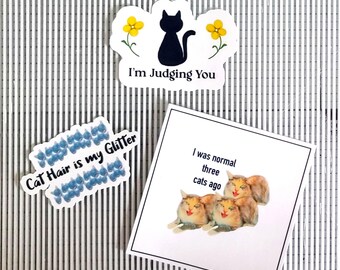 May include: Three stickers with cat-themed designs. One sticker features a black cat with yellow flowers and the text "I'm Judging You". Another sticker has the text "Cat Hair is my Glitter" with a pattern of blue cat paw prints. The third sticker shows three cats with the text "I was normal three cats ago".