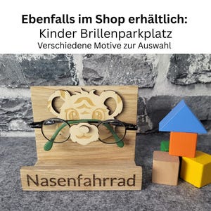 May include: Wooden eyeglasses holder with a bear design, holding a pair of green-framed glasses. The base of the holder is engraved with the word "Nasenfahrrad". Colorful wooden blocks are stacked to the right. Text at the top reads "Ebenfalls im Shop erh&auml;ltlich: Kinder Brillenparkplatz Verschiedene Motive zur Auswahl".