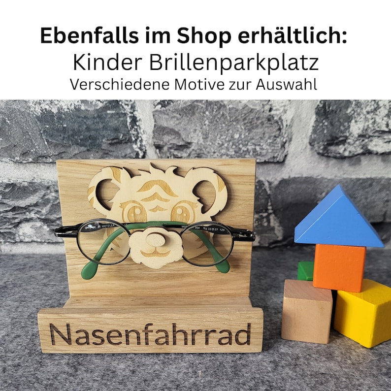 May include: Wooden eyeglasses holder with a bear design, holding a pair of green-framed glasses. The base of the holder is engraved with the word "Nasenfahrrad". Colorful wooden blocks are stacked to the right. Text at the top reads "Ebenfalls im Shop erh&auml;ltlich: Kinder Brillenparkplatz Verschiedene Motive zur Auswahl".