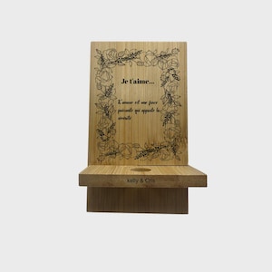 Peut inclure: Un support de téléphone en bambou avec un motif floral et le texte "Je t'aime..." en noir. Le support a une base rectangulaire et un dos avec une bordure florale. Le texte "L'amour est une force puissante qui apporte la sérénité" est également présent.