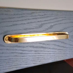 May include: A gold-colored, rectangular towel bar with rounded ends. The bar is mounted on a gray wooden surface. The bar has a polished, reflective surface, and the interior has a warm, golden glow.