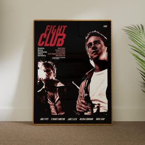 May include: A framed poster for the film "Fight Club" with a black background and red text. The poster features images of Brad Pitt and Edward Norton. The poster is displayed in a room with a plant.