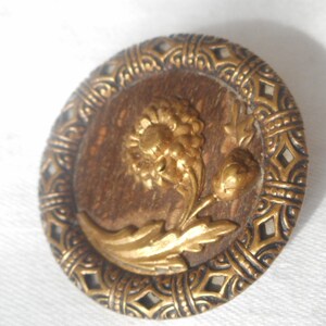 May include: A round, gold-toned button with a raised, detailed design of a flower with leaves. The button has a brown background and a decorative, patterned border.