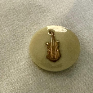 May include: A round, light green button with a gold-colored violin design. The button is slightly raised and has a glossy finish. The violin is detailed and centered on the button.
