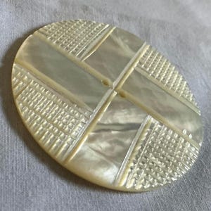May include: A round, off-white mother-of-pearl button featuring a geometric design. The surface is divided into four quadrants by intersecting lines, with alternating sections of smooth, iridescent shell and a textured, grid-like pattern.