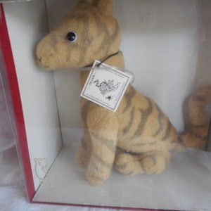 May include: A plush tiger toy in a box. The tiger is sitting up with its tail curled around its legs. The toy is brown and orange with black stripes. The box is white with a red border. The tag on the toy reads "Trigger - A John Wright & Co. Collectible".