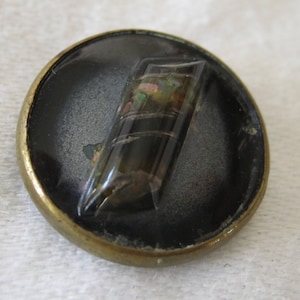 May include: A round, brass-colored button with a black background and a clear, iridescent, rectangular shape in the center.