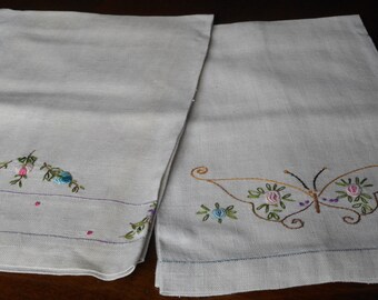 May include: Two white linen tea towels with embroidered floral designs. One towel features a butterfly with pink, green, and purple flowers. The other towel has a row of three blue flowers with pink centers.