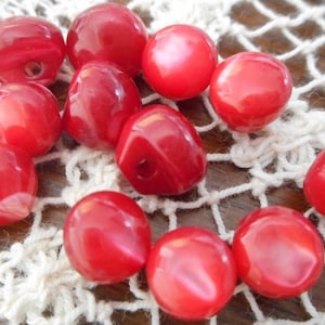 May include: A collection of 12 red, round, and slightly irregular shaped buttons. The buttons are made of a shiny, smooth material and have a hole in the center.