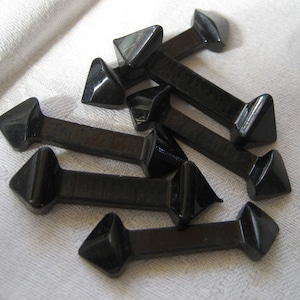 May include: Eight black glass arrow-shaped buttons with a pointed tip and a flat back. The buttons are arranged in a random pattern on a white surface.