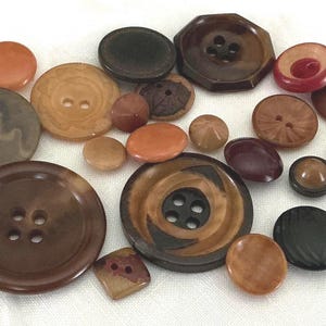 May include: A collection of various buttons in shades of brown, orange, and red. The buttons vary in size and shape, including round, oval, and geometric designs. Some buttons have two or four holes, while others have a shank. The buttons are made of different materials.