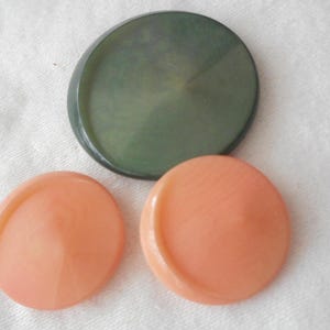 May include: Three buttons, one green and two peach colored. The green button is round and has a mottled surface. The peach buttons are round and have a smooth surface.