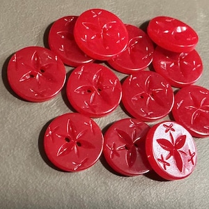 May include: A collection of red, round buttons with a floral design. Each button has two holes for sewing and a raised, stylized flower pattern. The buttons are made of a glossy, translucent material.